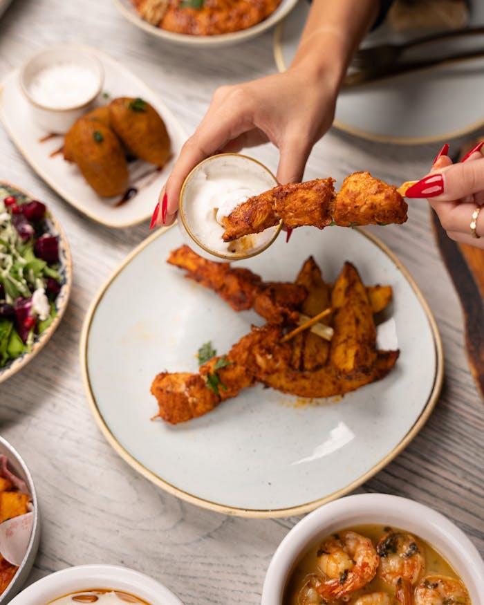 Close-up of chicken skewers being dipped in creamy sauce amidst a variety of dishes.